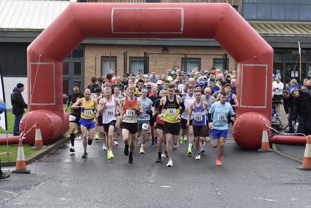 Start of a 10K race, large group of runners with numbers running under an inflatable red arch with a building in the background.
