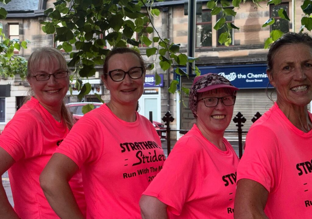 Four female runners standing in a line, side on with hands on hips and wearing bright pink running t-shirts.