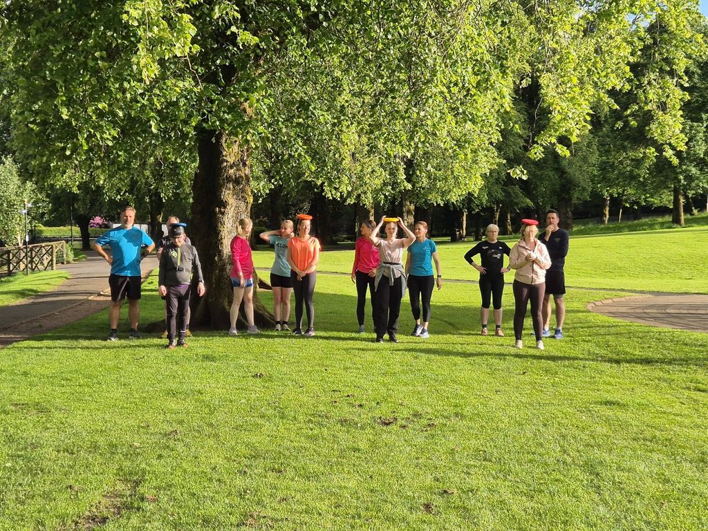 Group of runners in a park in front of a large tree, standing in small groups with one of each group with a coloured 'halo' for a sports day event