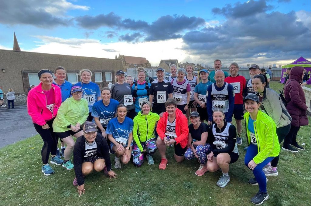 Group of runners standing and crouched, some are wearing race numbers, buildings and clouds in the background.
