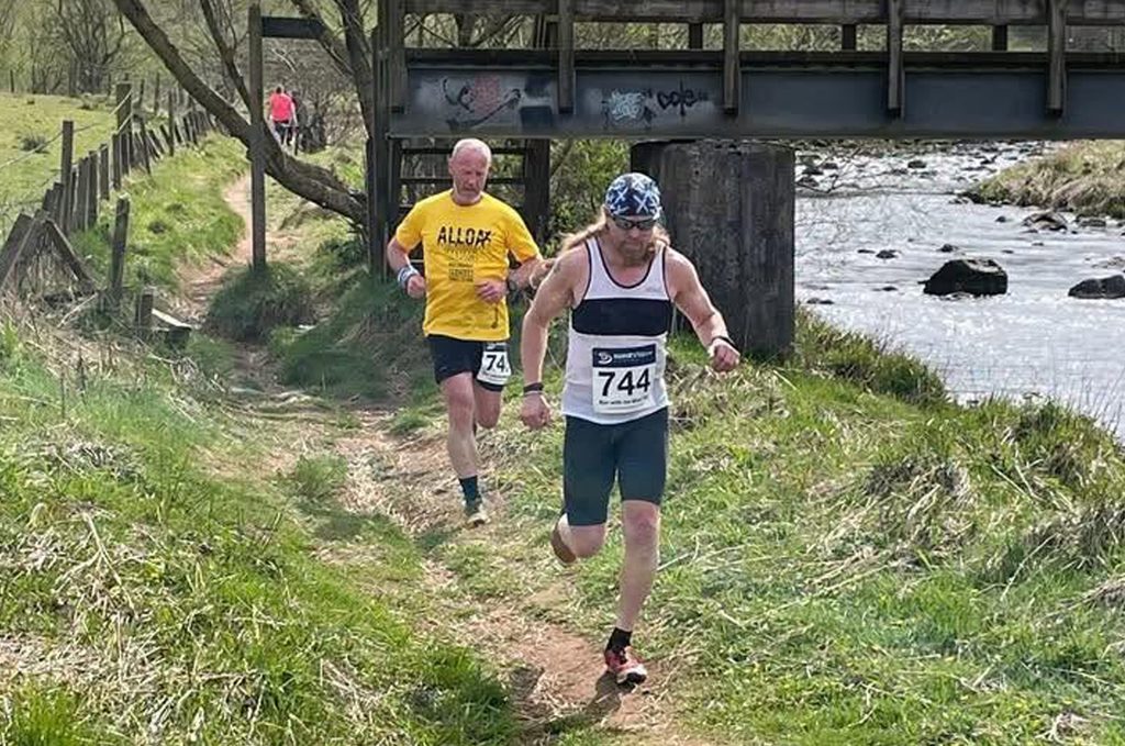 Two mail runners wearing race numbers running along a trail next to a river with a bridge in the background.