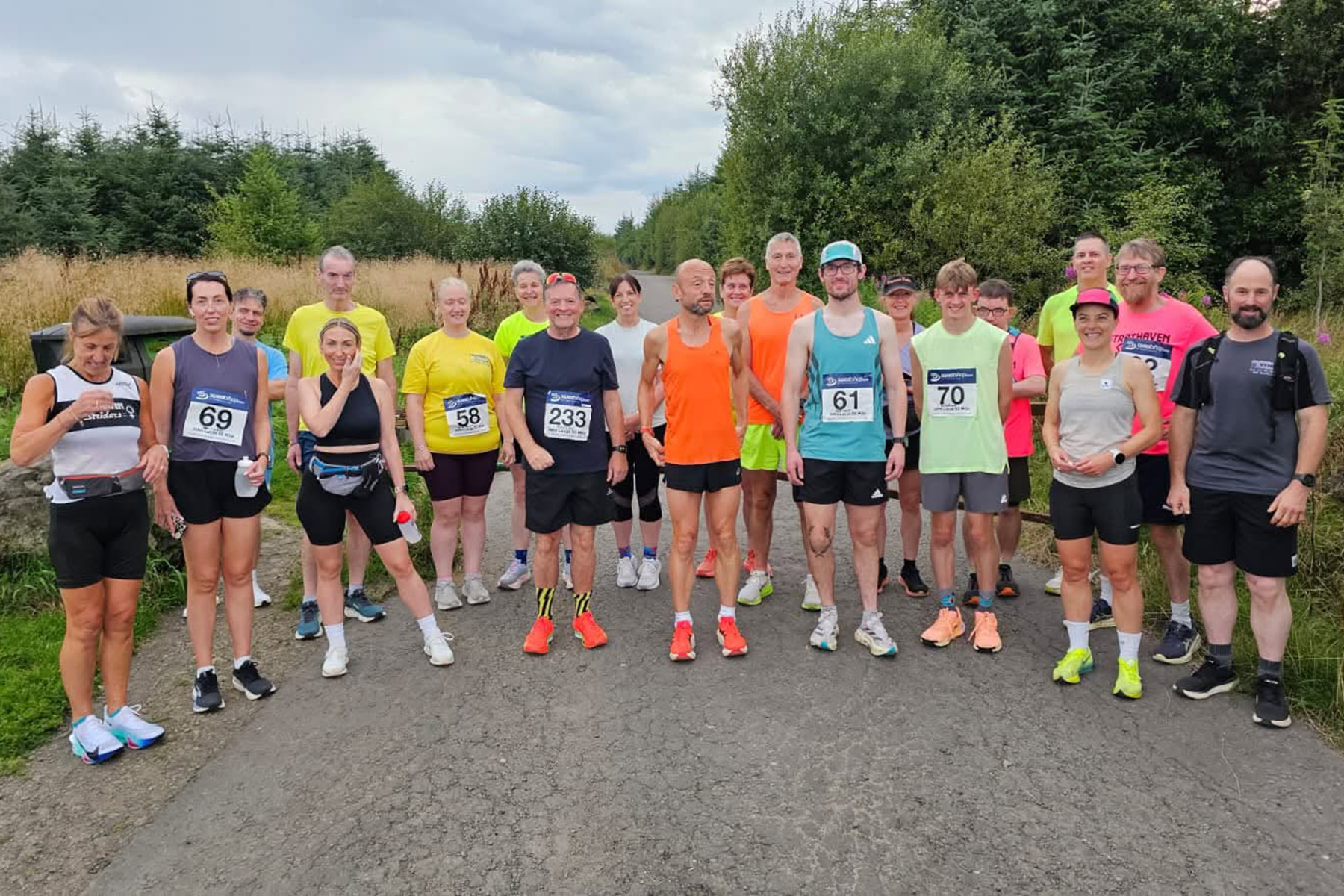 Group of male and female runners in running kit with race numbers, standing on a road in the countryside.